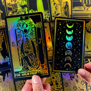 May include: Two tarot cards with a black background and gold foil accents. One card features "THE HIGH PRIESTESS" text and a seated figure. The other card shows the phases of the moon. Both cards have a holographic sheen.
