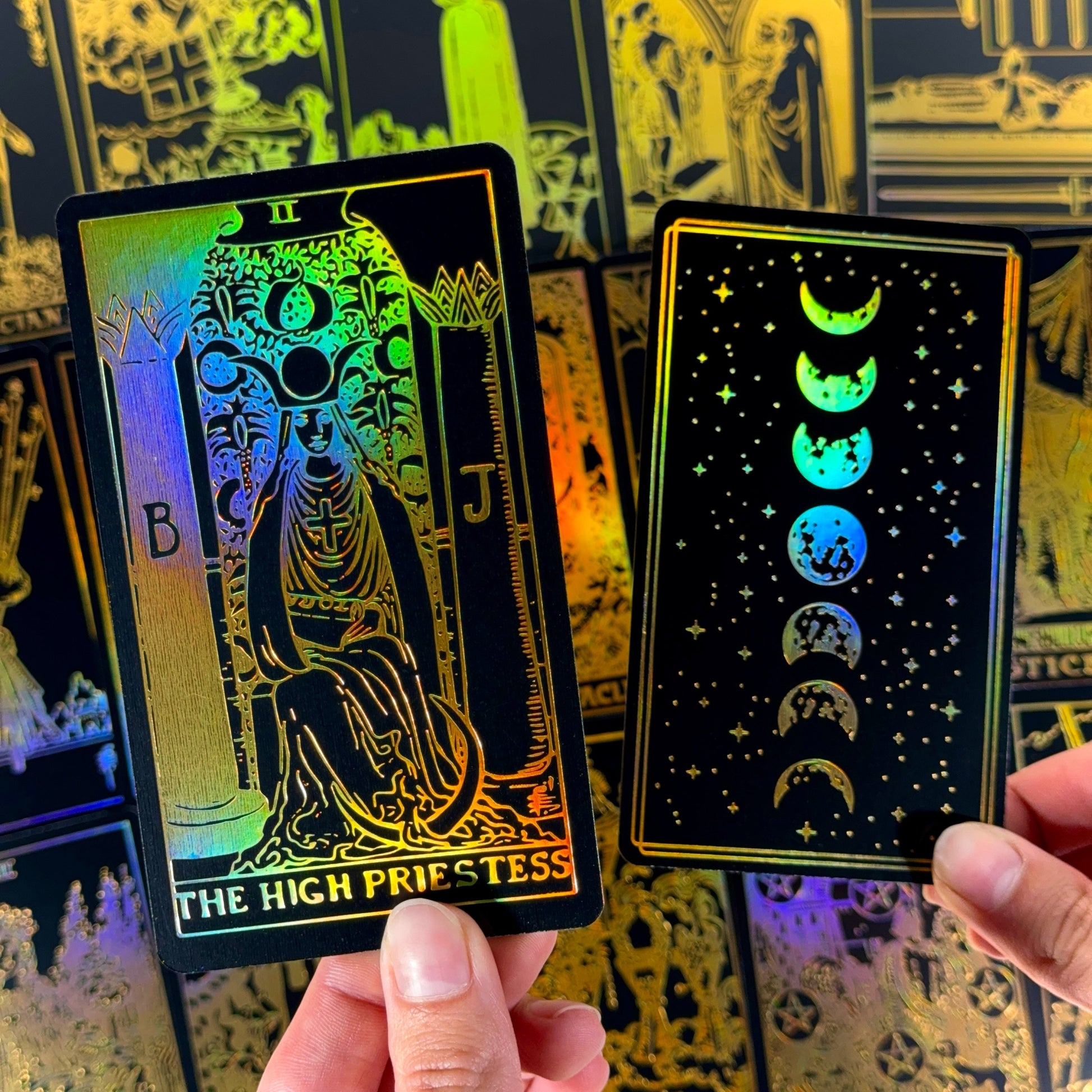 May include: Two tarot cards with a black background and gold foil accents. One card features "THE HIGH PRIESTESS" text and a seated figure. The other card shows the phases of the moon. Both cards have a holographic sheen.