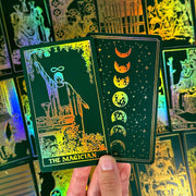May include: Two tarot cards with a dark green background and gold foil accents. One card features "THE MAGICIAN" with an illustration of a figure. The other card shows phases of the moon. Other cards are visible in the background.