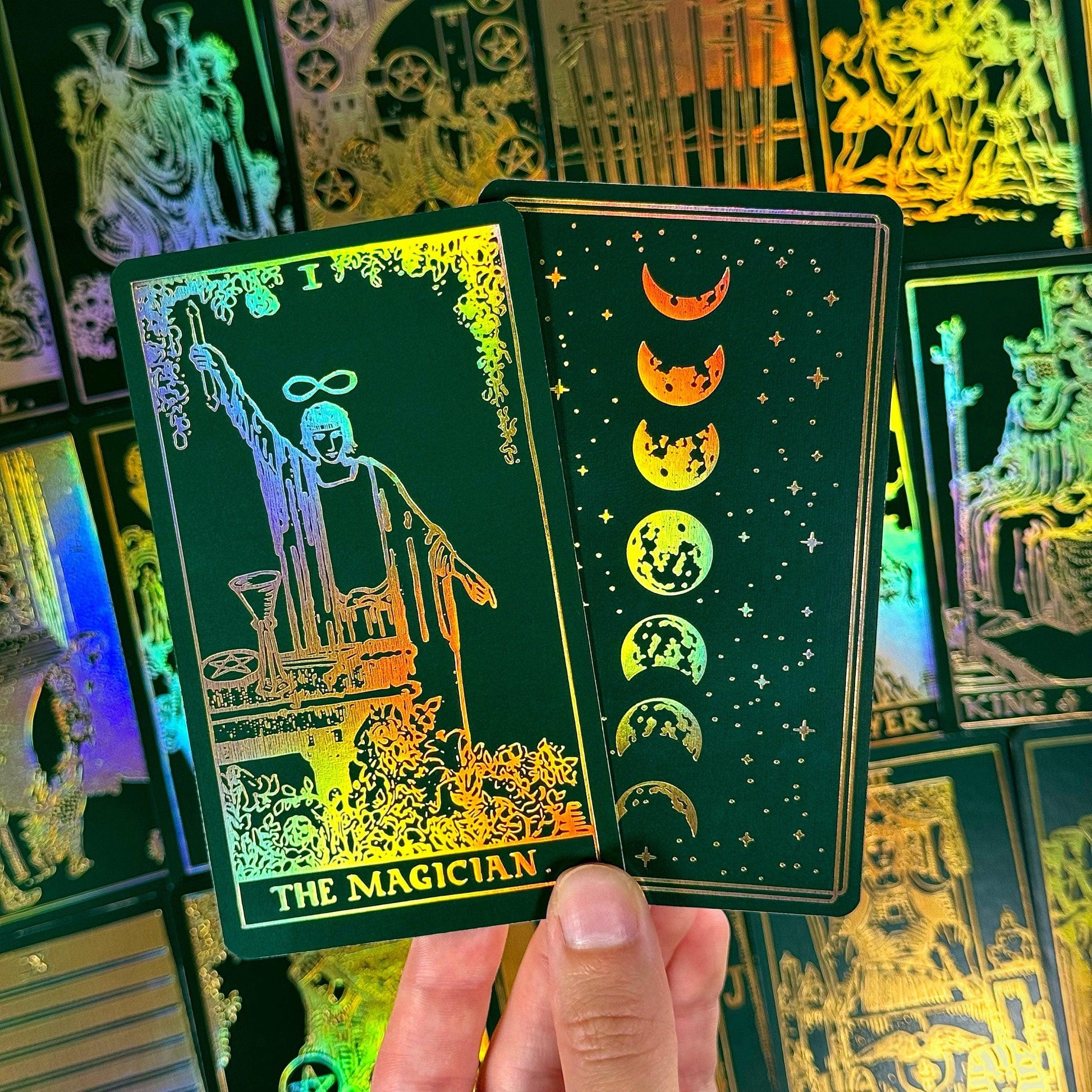 May include: Two tarot cards with a dark green background and gold foil accents. One card features "THE MAGICIAN" with an illustration of a figure. The other card shows phases of the moon. Other cards are visible in the background.